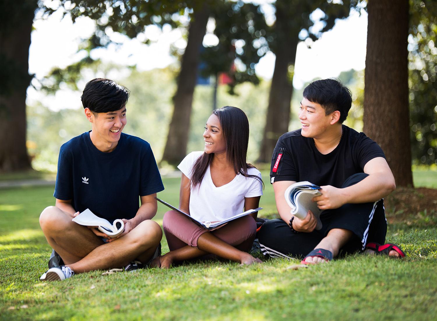 Three students sitting outside in the grass with textbooks.